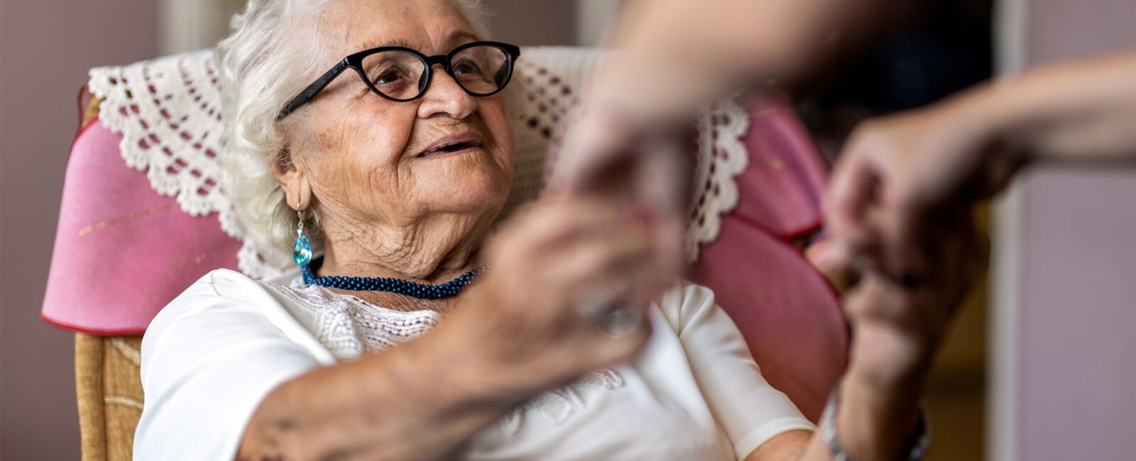 Elderly woman sitting in chair holding hands and smiling at her in-home caregiver.