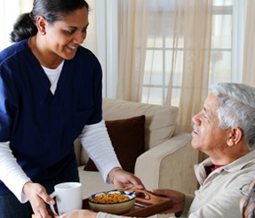 In-home caregiver helping client by giving him his breakfast in his living room.