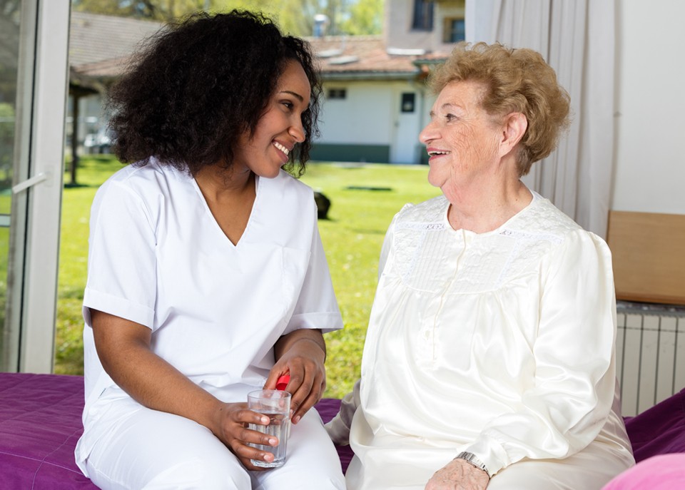Elderly woman and caregiver sitting near window with glass of water as caregiver provides medication reminder services.