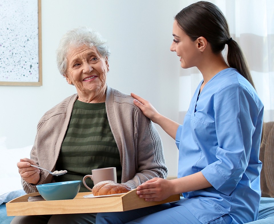 Young female caregiver in blue scrubs comforting elderly woman as she helps her with her meal.
