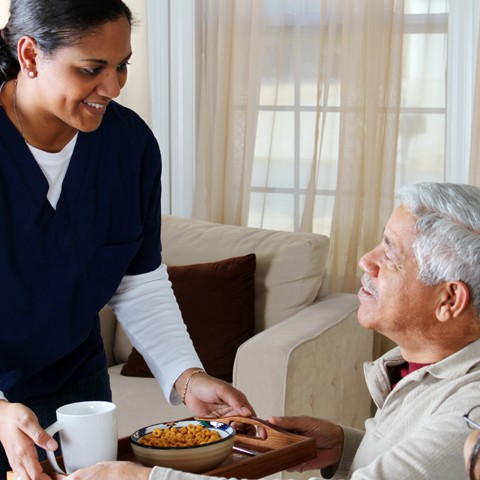 In-home caregiver helping client by giving him his breakfast in his living room.