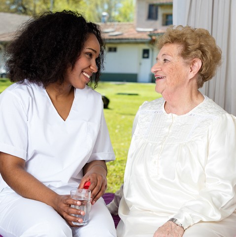 Elderly woman and caregiver sitting near window as caregiver reminds client to take medication.