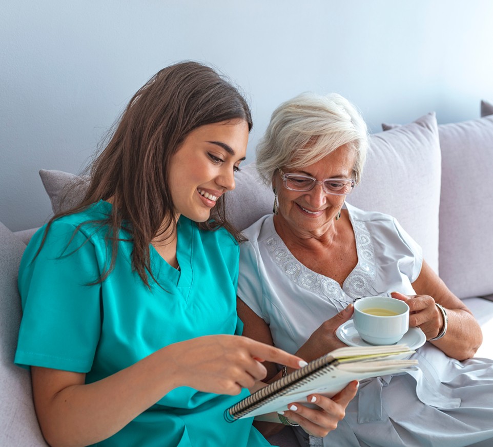 Young caregiver and elderly female read book together.