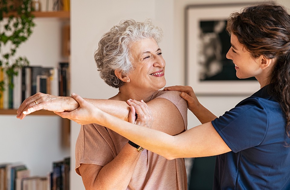 Image of elderly woman stretching her arm and a PT helping her.