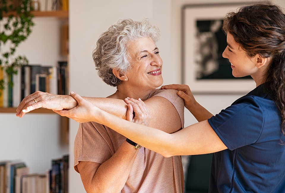 Image of elderly woman stretching her arm and a PT helping her.