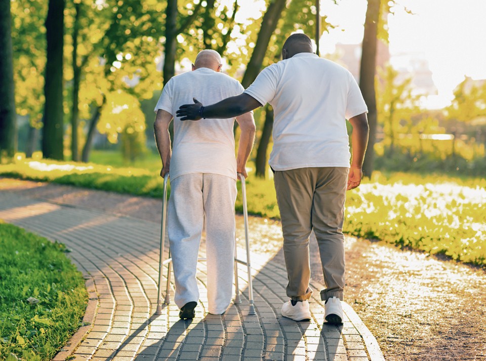 Photo from behind of male caregiver guiding his client outdoors while the client holds on to his walker.
