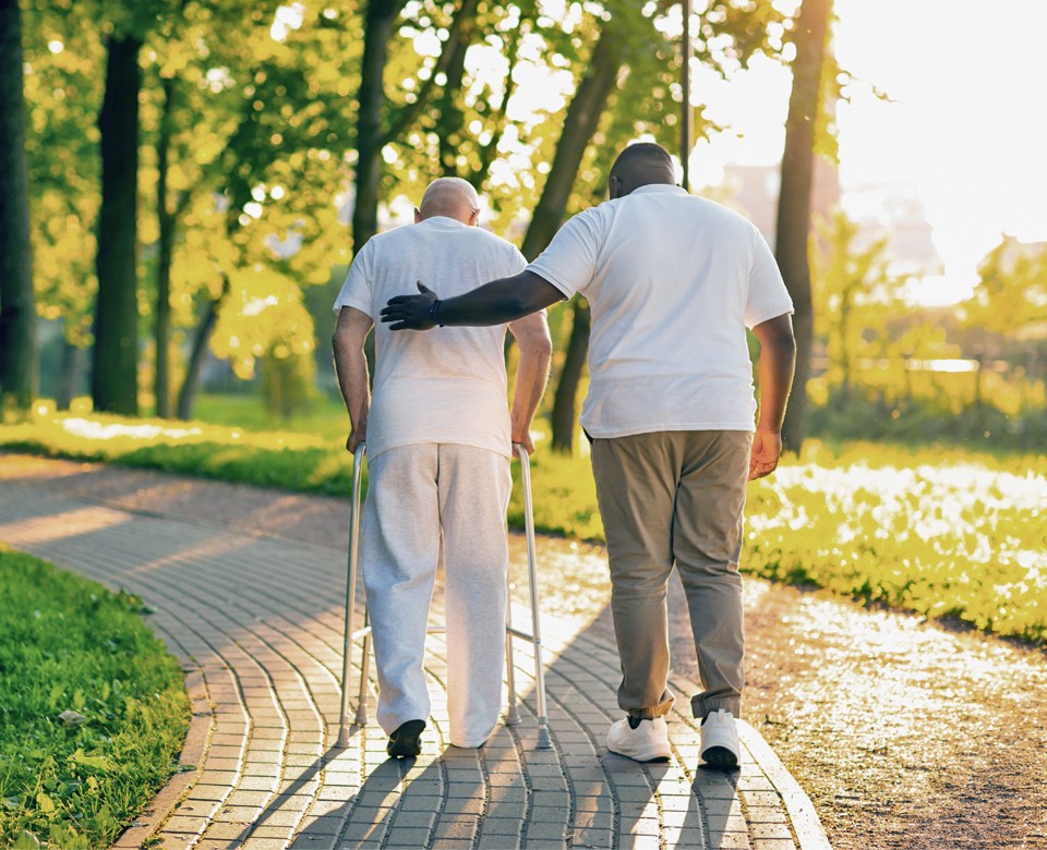 Photo from behind of male caregiver guiding his client outdoors while the client holds on to his walker.