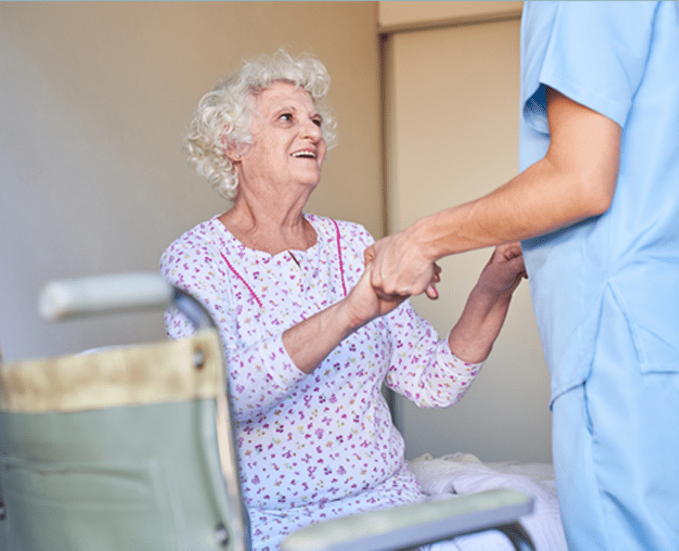 Male Health Care Worker Holds Hands with Eldery Woman Sitting on Her Bed